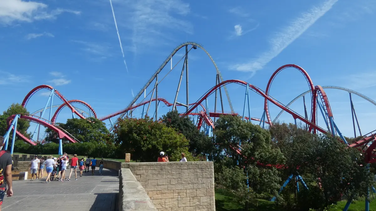 A red and blue steel roller coaster loops against a blue sky over a stone wall and a path with visitors walking