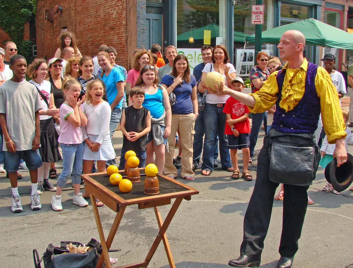 A street magician in a purple waistcoat holds an orange above a small folding table with more oranges on it, in front of a crowd of children and adults