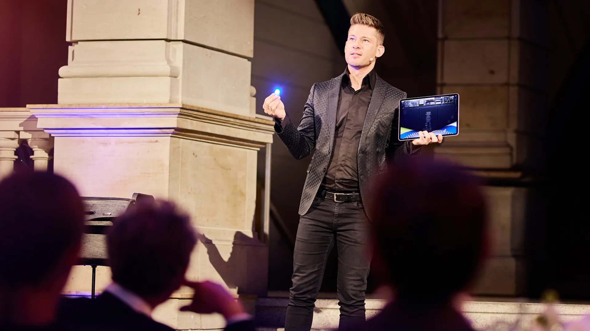 A stage magician in a dark patterned jacket holds a glowing blue light in one hand and a tablet in the other, with the heads of the audience in the foreground