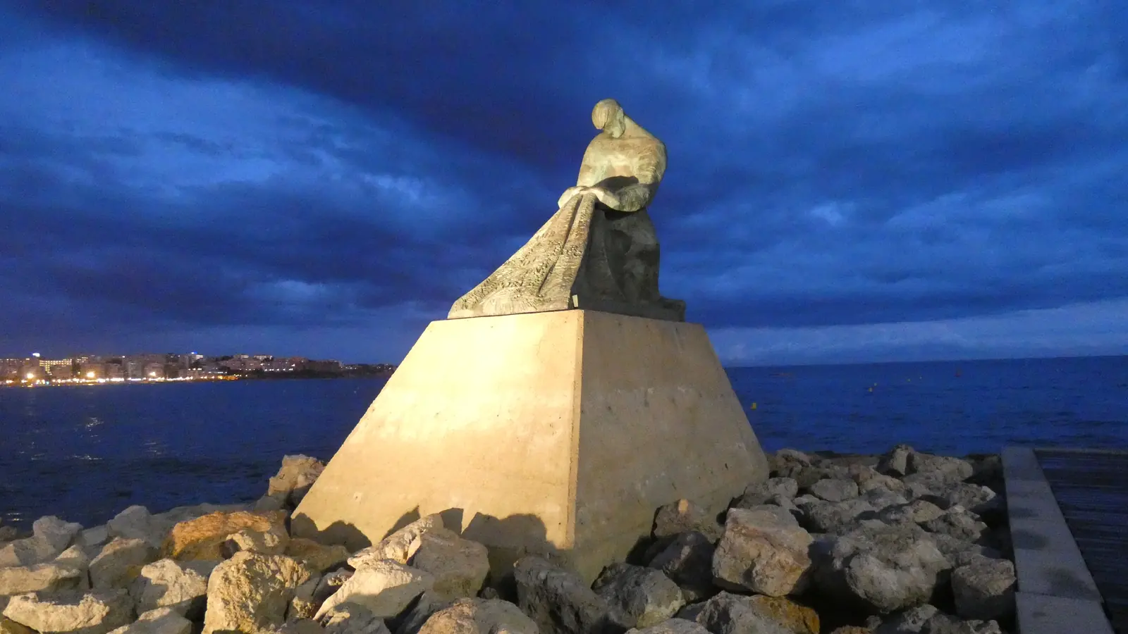 The Monument to Fishermen at Salou, a bronze figure on a stone pyramid on the rocks of the breakwater, with the lights of the town across the water under a deep blue dusk sky