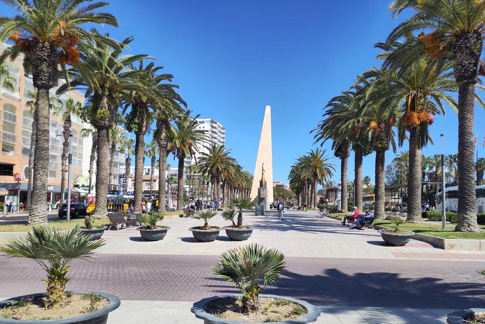 The Passeig Jaume I in Salou, a wide paved promenade lined with palm trees and planters, leading towards a tall slim obelisk